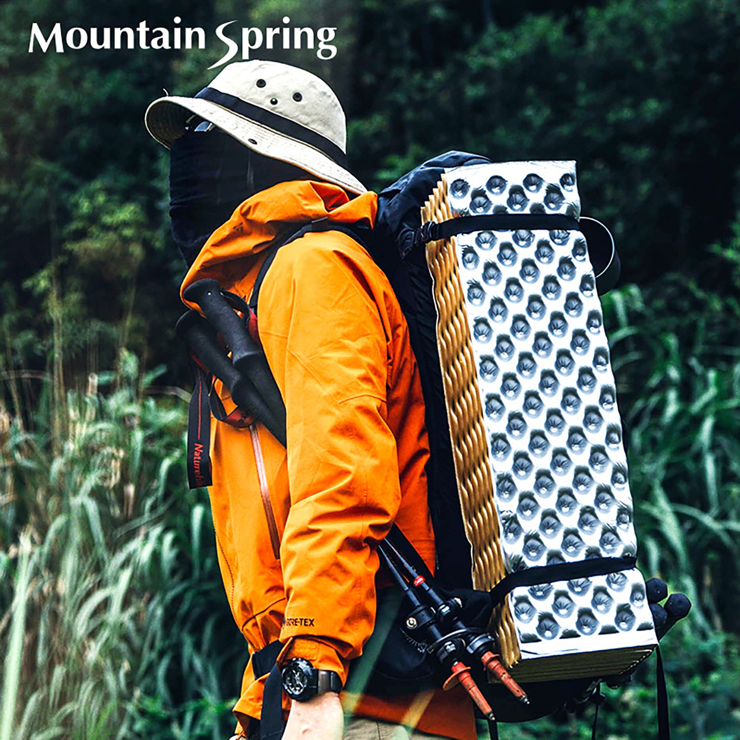 A person wearing an orange Gore-Tex hard-shell jacket, a bucket hat, and sunglasses carried a hiking backpack with a Mountain Spring folding foam pad tied to the side of the backpack.