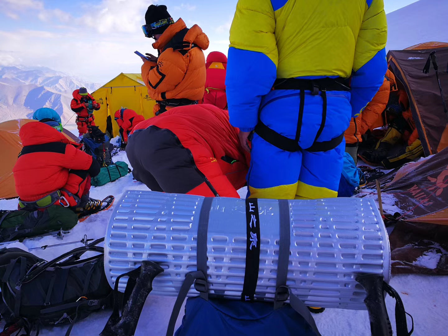 At the snow mountain base camp, many people in professional mountaineering gear are resting in the background, while in the center of the image is a roll-up Mountain Spring reflective foam pad.