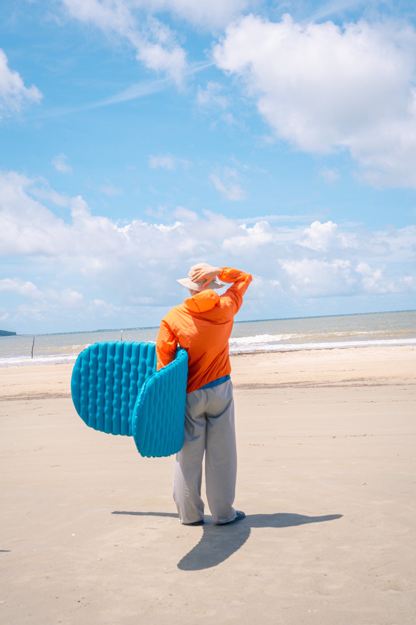 On the beach, the wind is very strong, and a man in an orange windbreaker and a khaki fishing hat is holding a hat with a blue R2M inflatable mat in his arms.