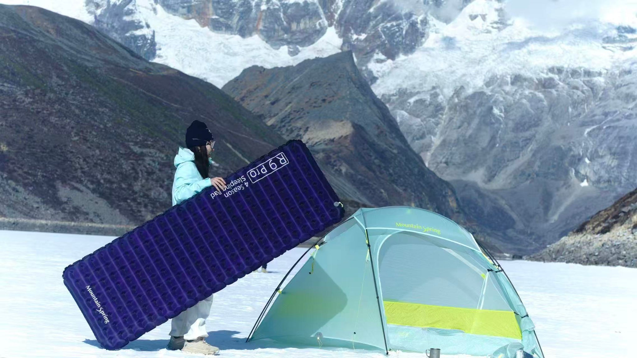 A woman in a blue down jacket and a black hat stands next to a light cyan DuFeng Pro tent, holding a purple R9 Pro inflatable air pad, in the snow, with a magnificent mountain of snow in the distance.