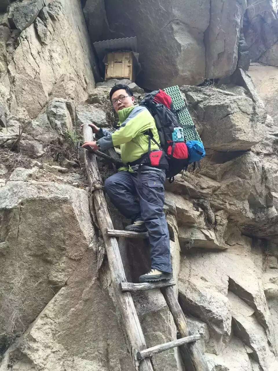 Mr. Cao, one of the founders of Mountain Spring, is carrying a hiking backpack and wearing a green jacket. He stands on a ladder that leans against a near-vertical rock face.